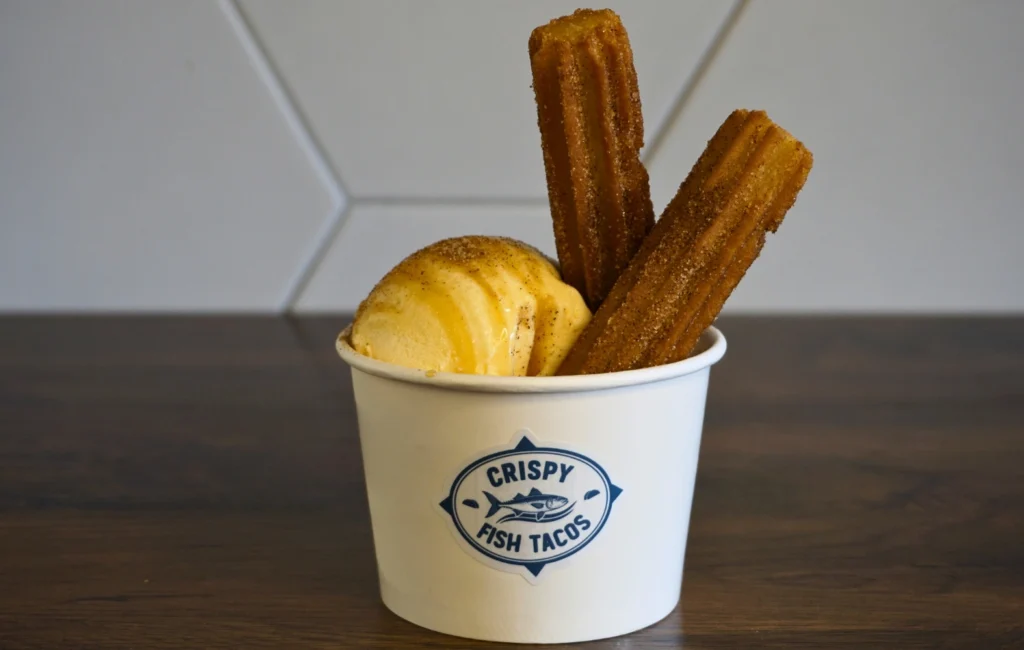 A cup filled with churros and a scoop of ice cream, topped with chocolate sauce and a sprinkle of nuts.