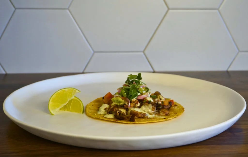 A taco on a white plate accompanied by a lime wedge, showcasing vibrant colors and fresh ingredients.